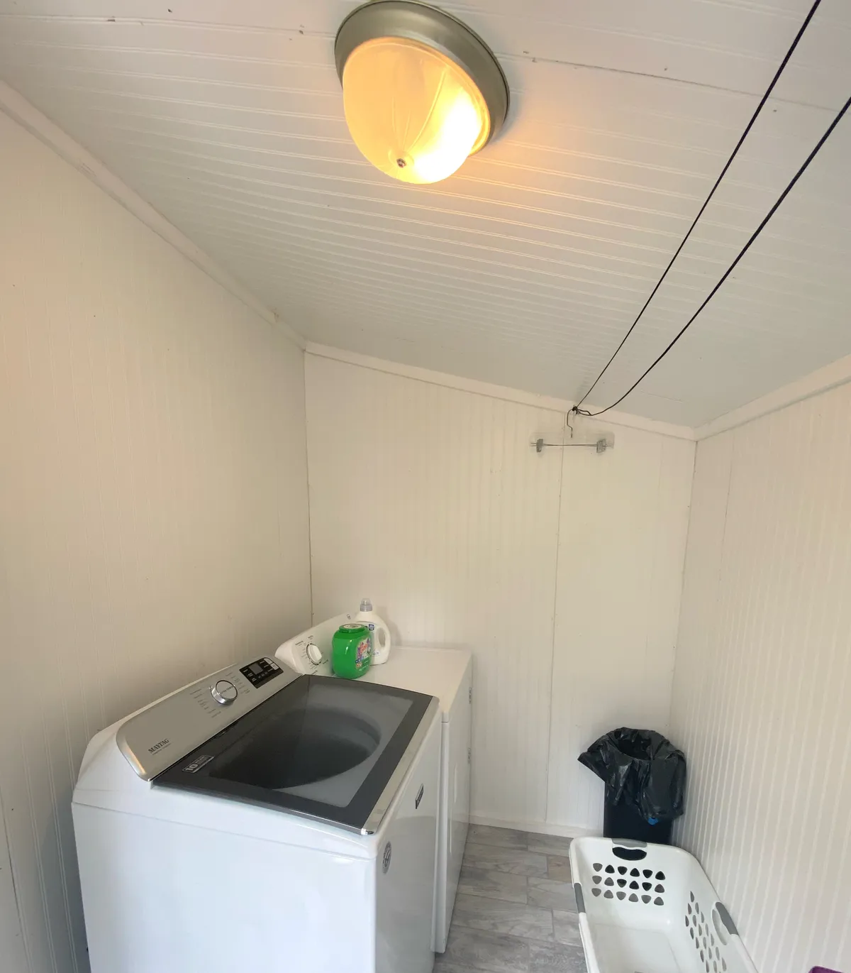 This image shows the laundry room with a washing machine, laundry basket, and a green detergent bottle. The environment is practical and functional, emphasizing utility and convenience for guests needing to do laundry during their stay.