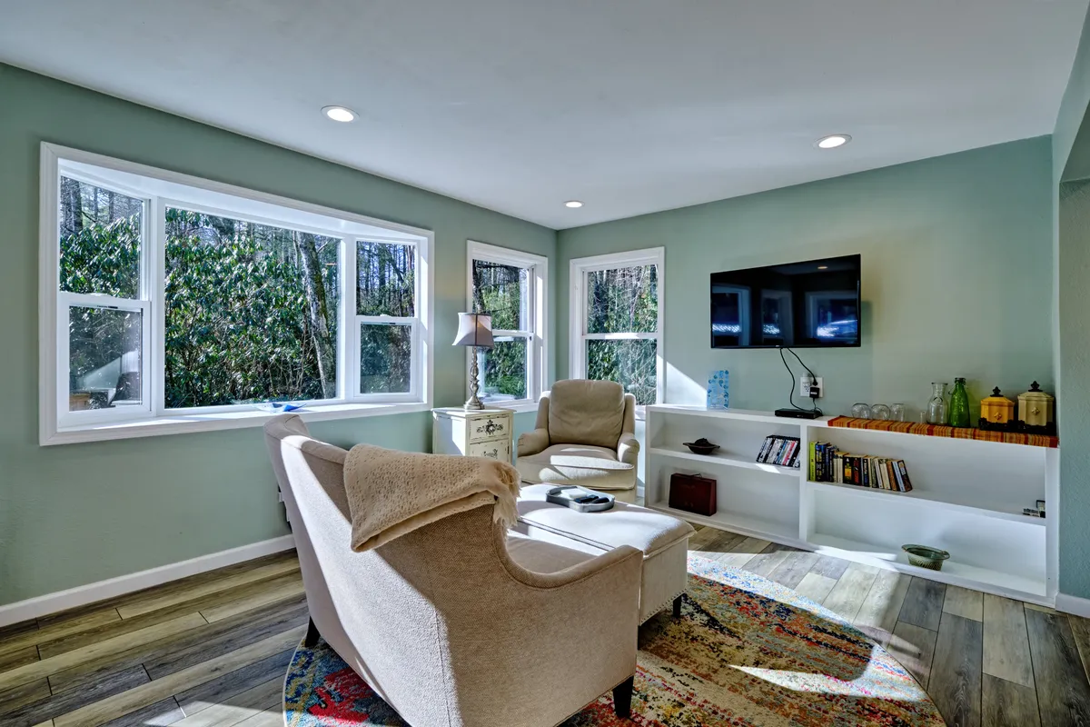 This image presents the living area featuring large windows that fill the room with natural light. Comfortable chairs are arranged for both relaxing and socializing, while a streaming-capable TV and a decorative lamp add to the cozy ambiance. The windows offer a glimpse of outdoor greenery, hinting at scenic views beyond.