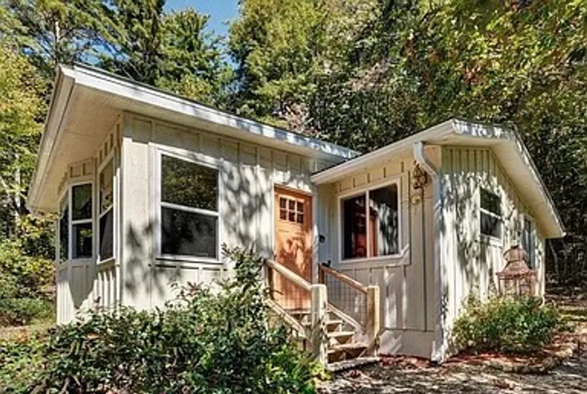 The image showcases the charming front entrance of a renovated bungalow, framed by large windows that let natural light spill inside. The cottage is nestled amid lush greenery, offering a tranquil setting with inviting porch seating. You can see the reflection of the nearby pond and hints of a firepit area, suggesting relaxing evenings outdoors. The overall atmosphere is peaceful and welcoming, with the cottage blending seamlessly into its serene, natural surroundings.