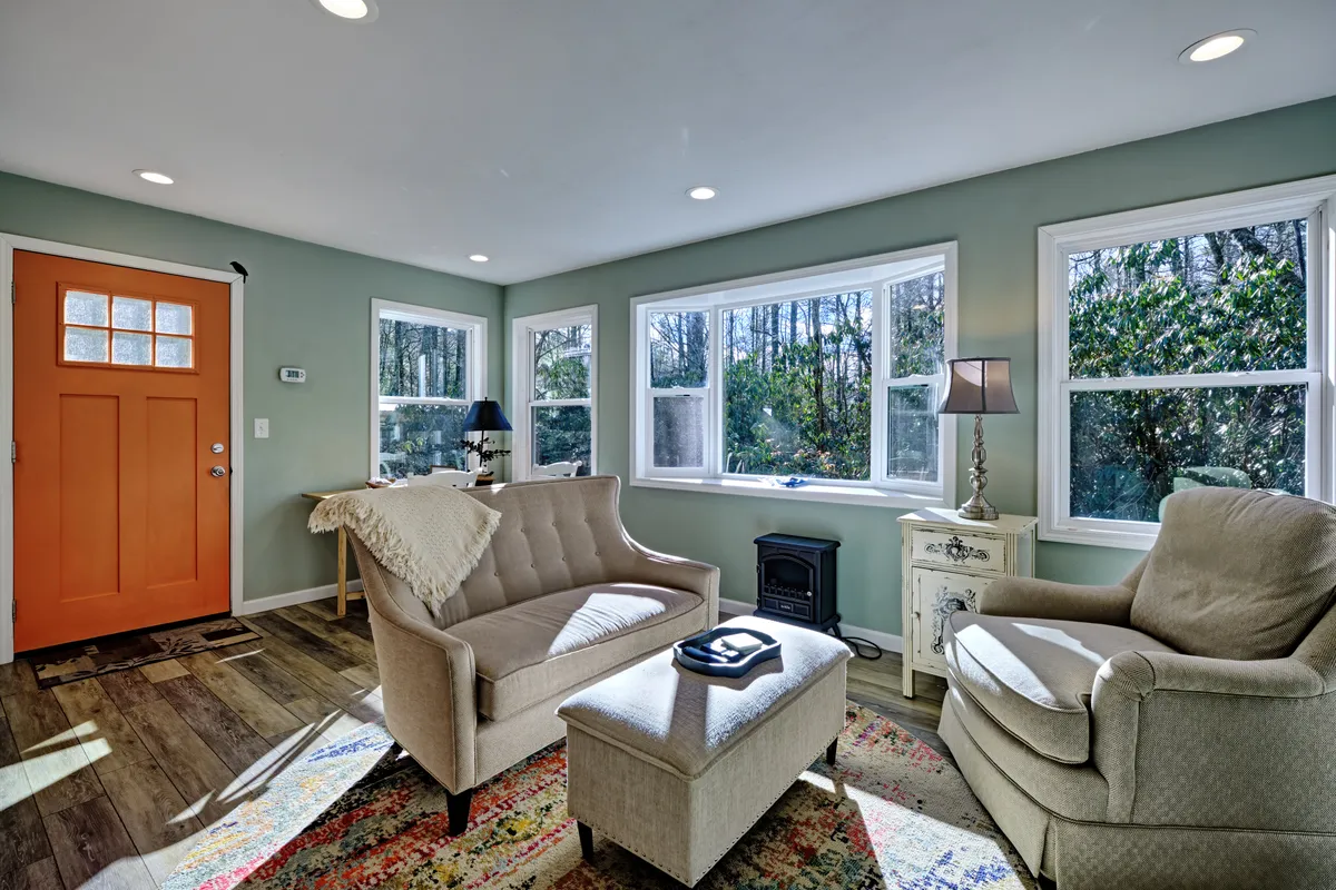 A welcoming corner of the living space with a vibrant orange front door, windows revealing lush greenery outside, and comfortable seating arranged for cozy gatherings. Sunlight streams through the windows, creating a warm and inviting atmosphere ideal for reading or enjoying coffee.