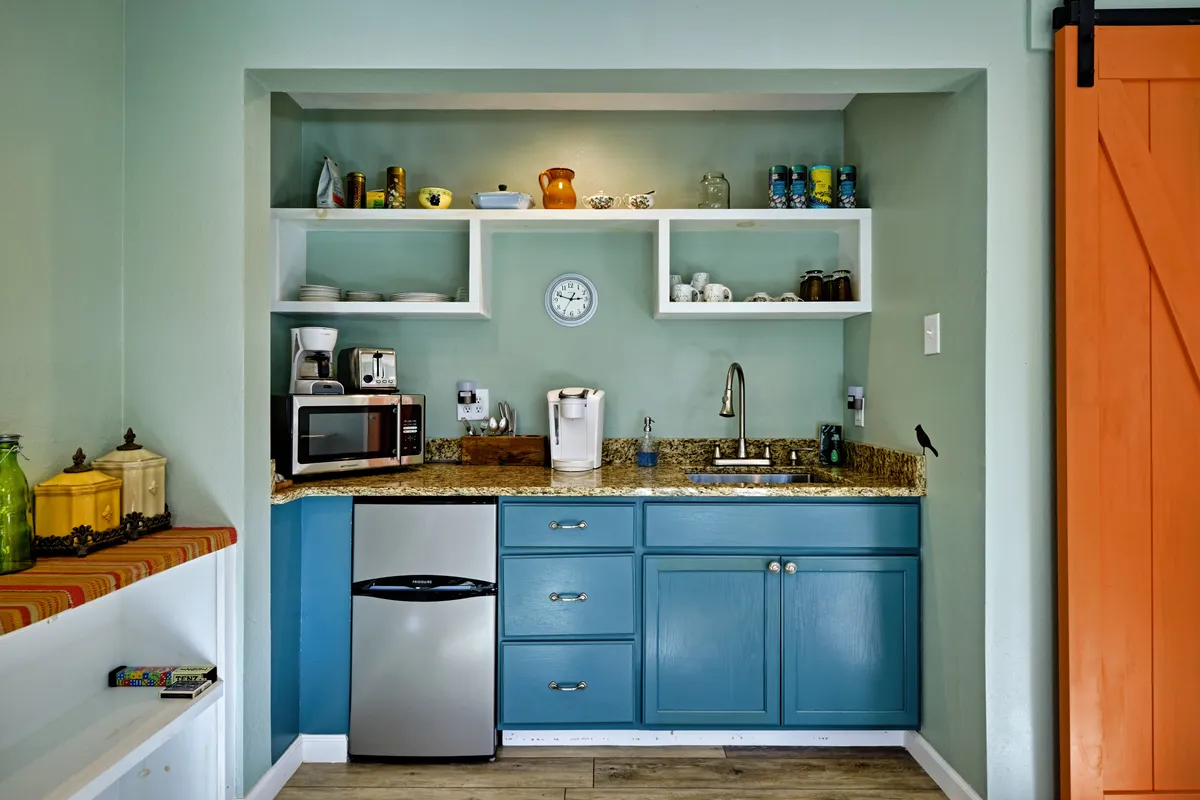 This image presents a close-up view of the kitchenette, focusing on blue cabinetry, a granite countertop, and open shelves stocked with glassware. The vignette suggests a well-equipped, attractive space ideal for preparing coffee, breakfast, or snacks. The overall mood is casual and welcoming, with a lived-in feel.