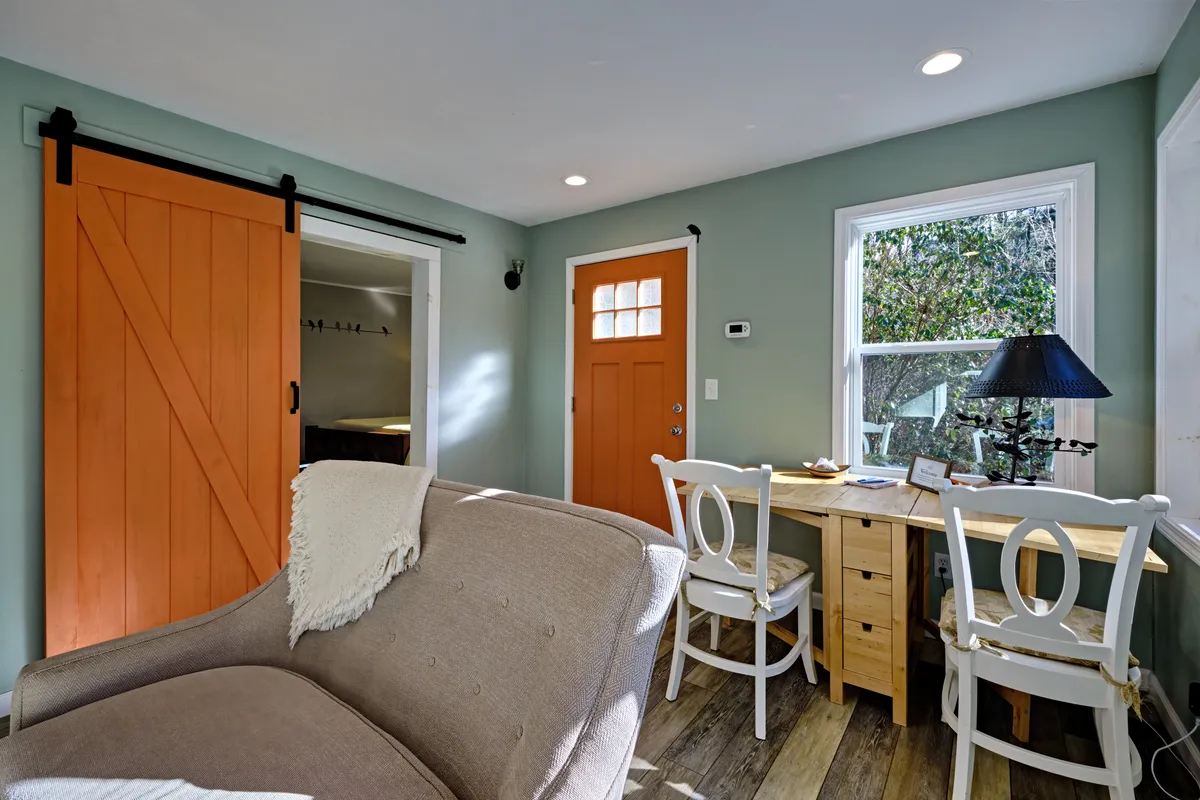 A welcoming interior scene featuring an orange barn door, a sturdy wooden table, and a comfortable armchair. Sunlight streams through a window, illuminating the cozy space, and casting warm tones across the room. The setup invites relaxation, reading, or enjoying a meal, with hints of the outdoors visible through the window.
