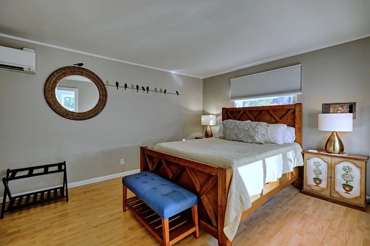 A tranquil bedroom with a neatly made bed featuring soft linens, flanked by two bedside lamps and adorned with tasteful artwork. A large decorative mirror adds depth to the space and natural light filters in, creating an inviting, restful ambiance.