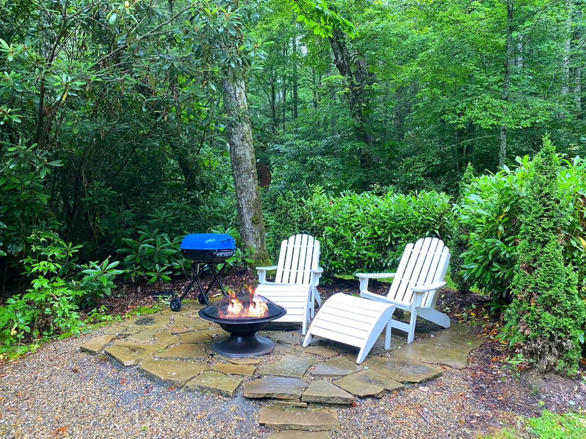 An inviting outdoor fire pit area surrounded by adirondack chairs, nestled in lush greenery. The scene is set for evening gatherings, with a grill nearby and space for guests to relax and enjoy the tranquil surroundings.