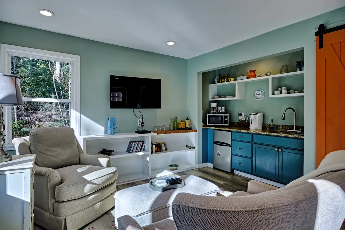 This image features a cozy interior with comfortable armchairs positioned near a kitchenette area, a television, and shelves adorned with decor. The kitchenette is visually accessible and equipped for light meal preparation, aligning with the section’s emphasis on practical essentials. The scene feels warm and welcoming, suggesting a space where guests can relax or enjoy snacks and meals.